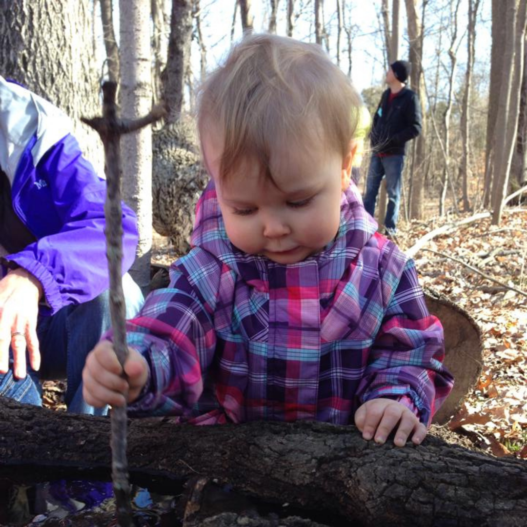 A toddler holds a stick vertically and is using it to poke a log. They are wearing a pink and purple plaid coat. 